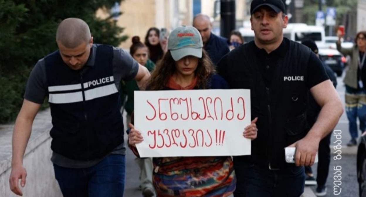 Полицейские ведут активистку, держащую плакат с надписью: "Анцухелидзе бессмертен!". Фото: Леван Зазадзе / Pirveli https://tvpirveli.ge/ka/siaxleebi/sazogadoeba/103394-aqtivisti-plakatit-antsukhelidze-ukvdavia-5000-larit-daajarimes